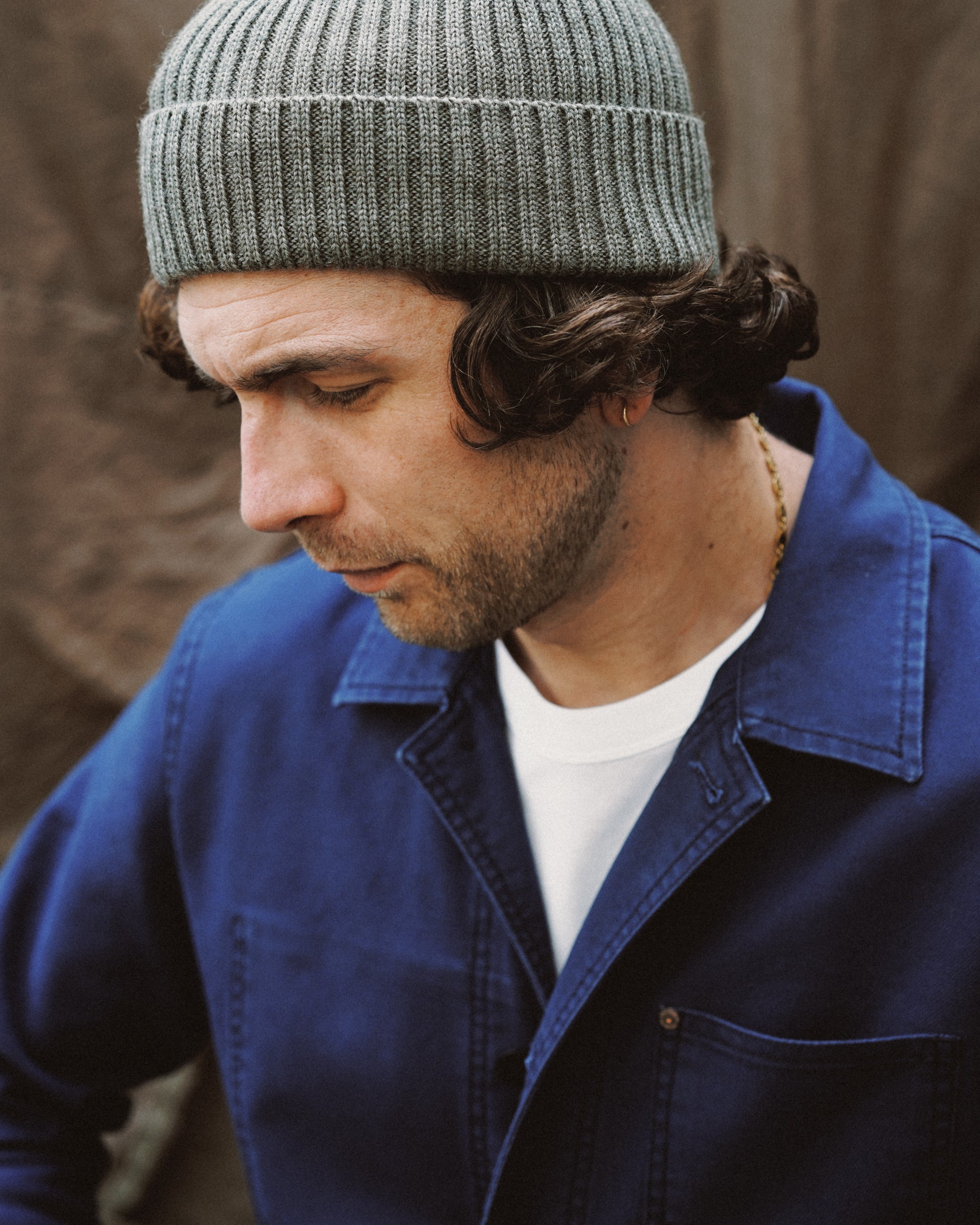 Un mannequin arbore une veste de travail vintage associée à un bonnet en laine kaki et un t-shirt blanc à col rond de la marque Champ de Manœuvres, dans un style d'inspiration militaire.