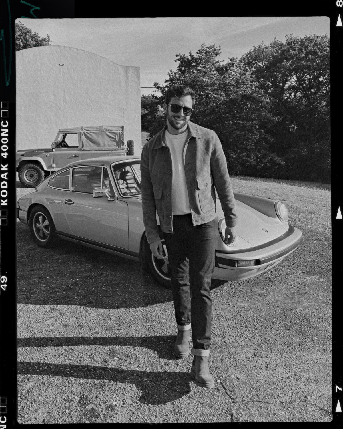 mannequin portant un blouson en cuir velours vintage devant une porche 911 et un landrover explorer militaire