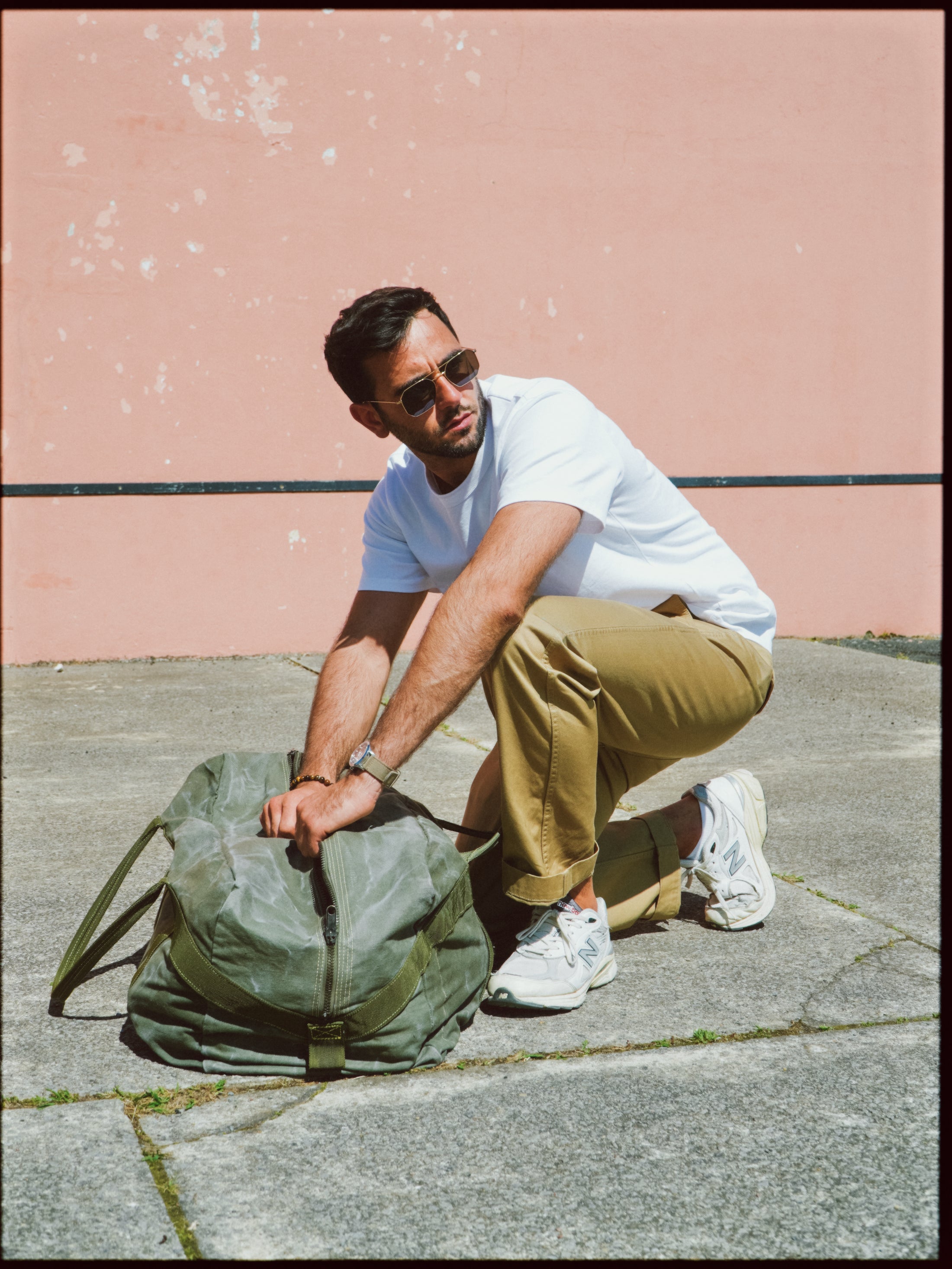Homme portant un t-shirt blanc, un pantalon chino sable et des baskets New Balance, accroupi sur un sol en béton tout en ouvrant un sac de voyage vert, contre un mur rose texturé.