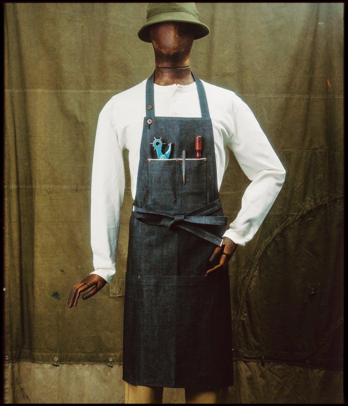 Tablier de métier en denim selvedge porté sur un mannequin avec un henley blanc, devant une toile de tente kaki