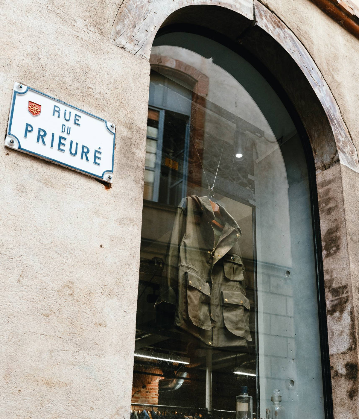 Plaque émaillée de la rue du Prieuré à Toulouse et veste field olive en vitrine de la boutique Champ de Manœuvres