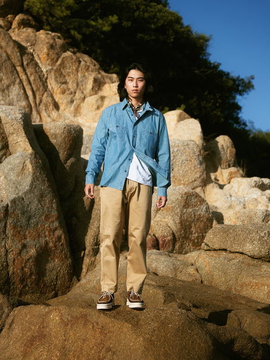 Tenue complète Champ de Manoeuvres avec chemise en denim bleu clair, pantalon beige chino et chaussures bateau, photographiée en extérieur sur des rochers"