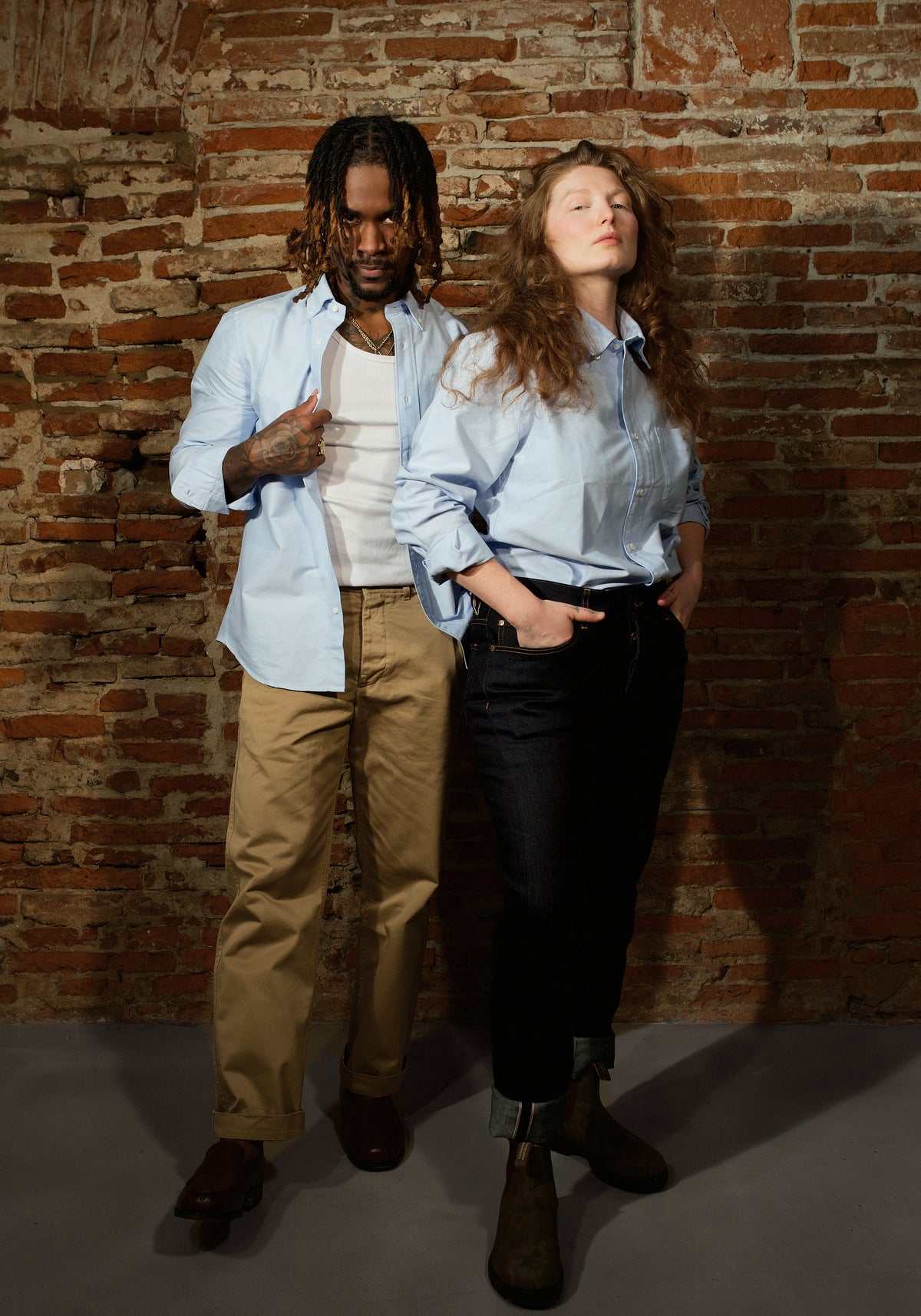 Deux mannequins en chemise oxford bleue devant un mur de brique, l'une ouverte sur un débardeur, l'autre manches retroussées