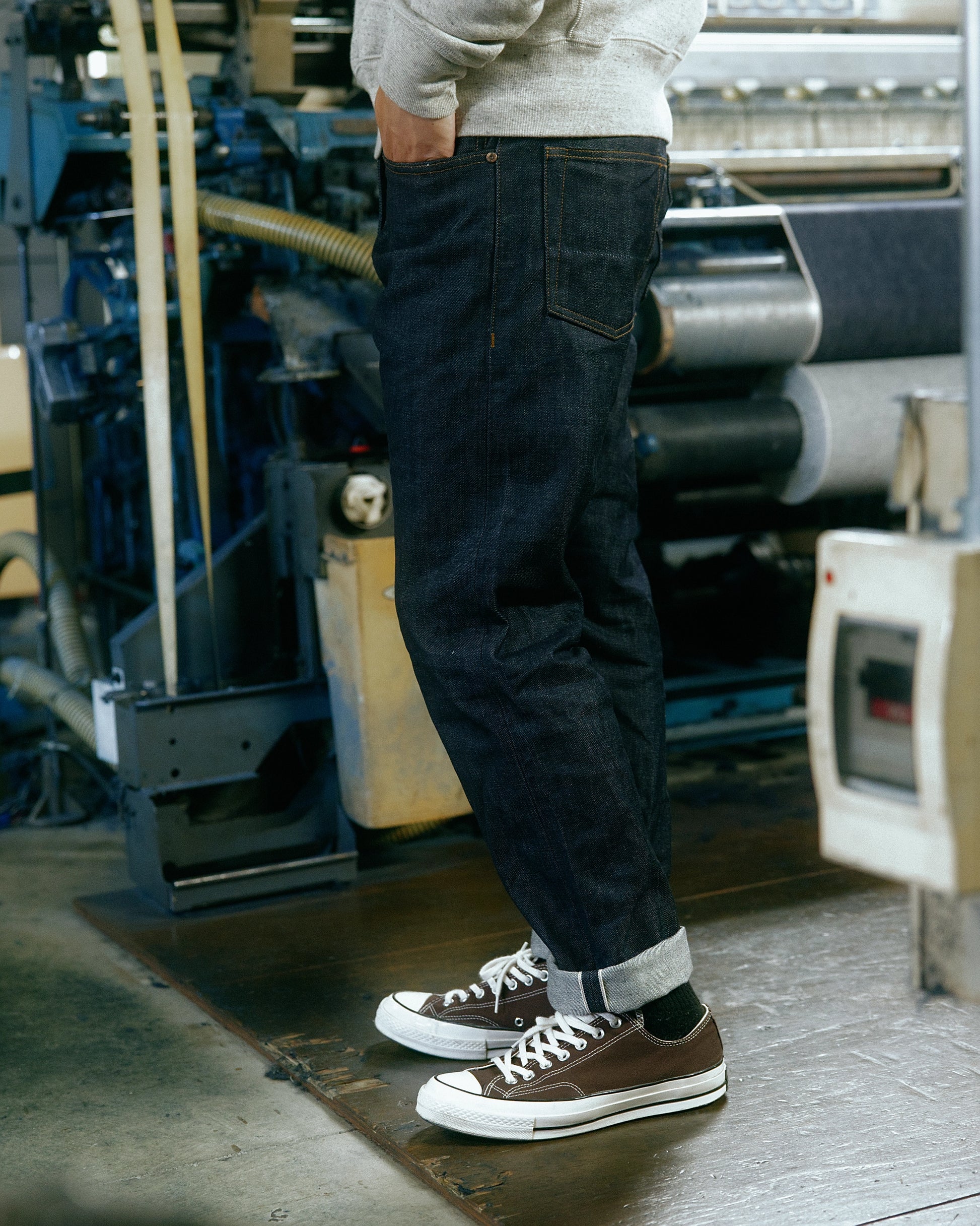 Détail d'un jean selvedge japonais porté avec des baskets, capturé dans une usine de tissage traditionnelle, symbolisant le savoir-faire du denim haut de gamme.