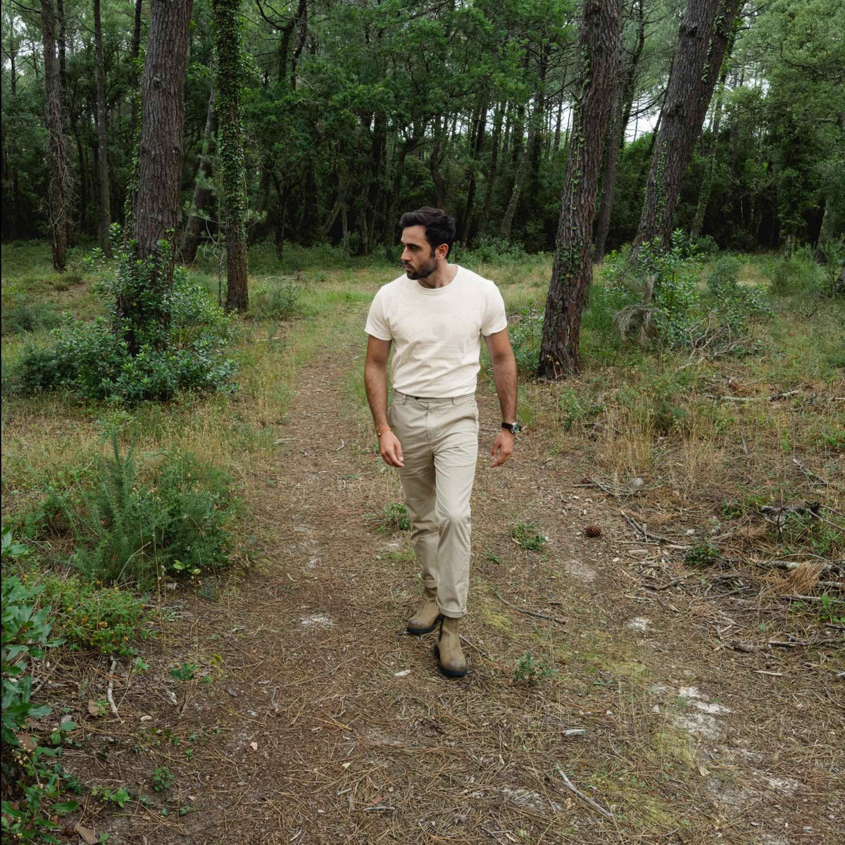 Homme marchant sur un sentier de forêt de pins, portant le t-shirt colletage écru et un pantalon beige