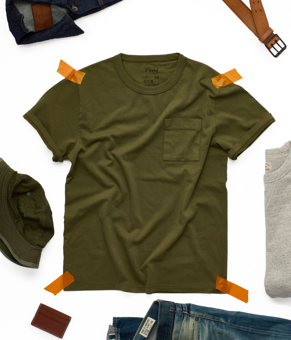 T-shirt kaki homme à poche posé à plat, entouré d'une veste en jean brut, d'une ceinture en cuir et d'un bob