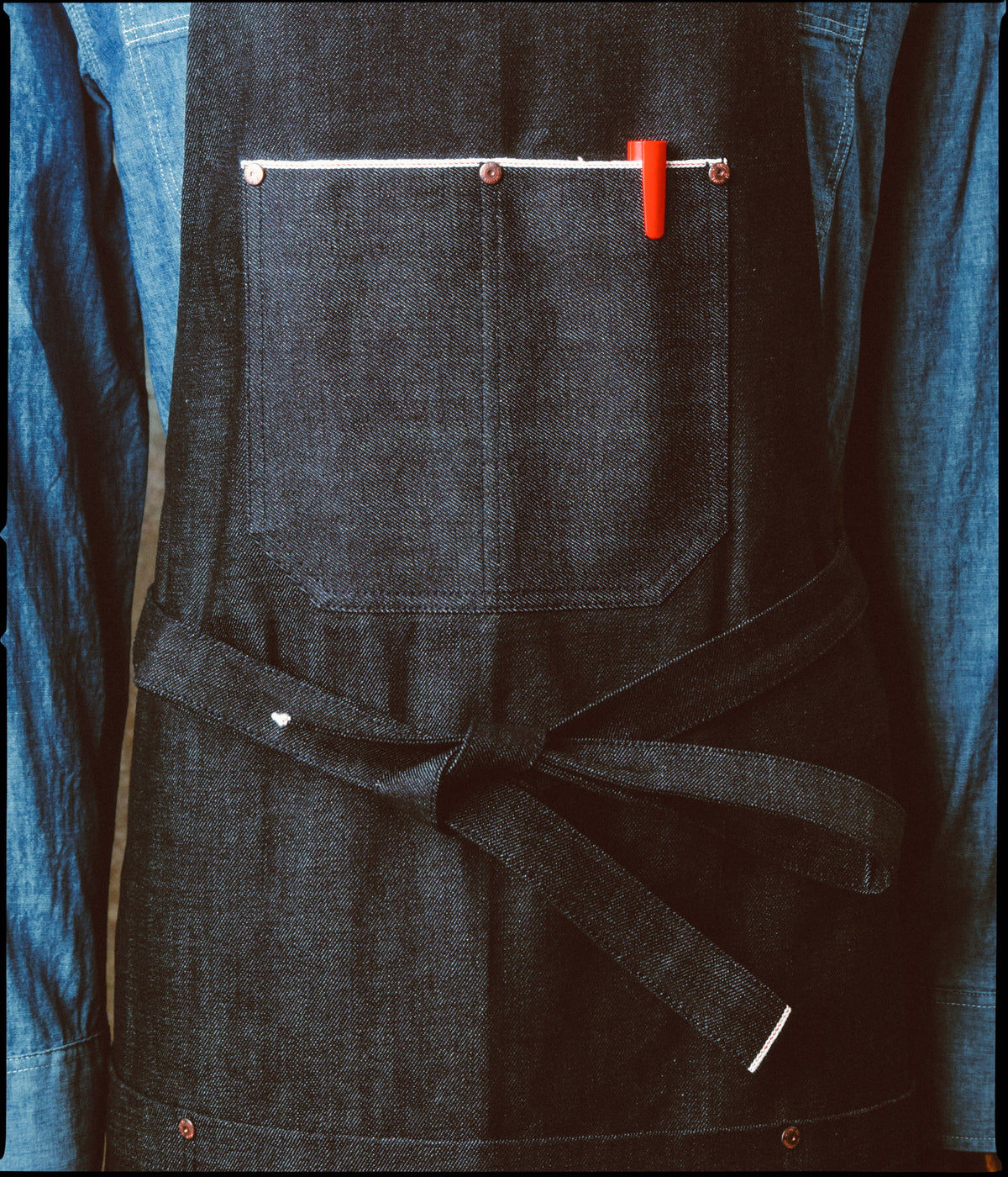 Détail des poches du Tablier de métier en denim selvedge et de son cordon de taille noué, sur une chemise en chambray