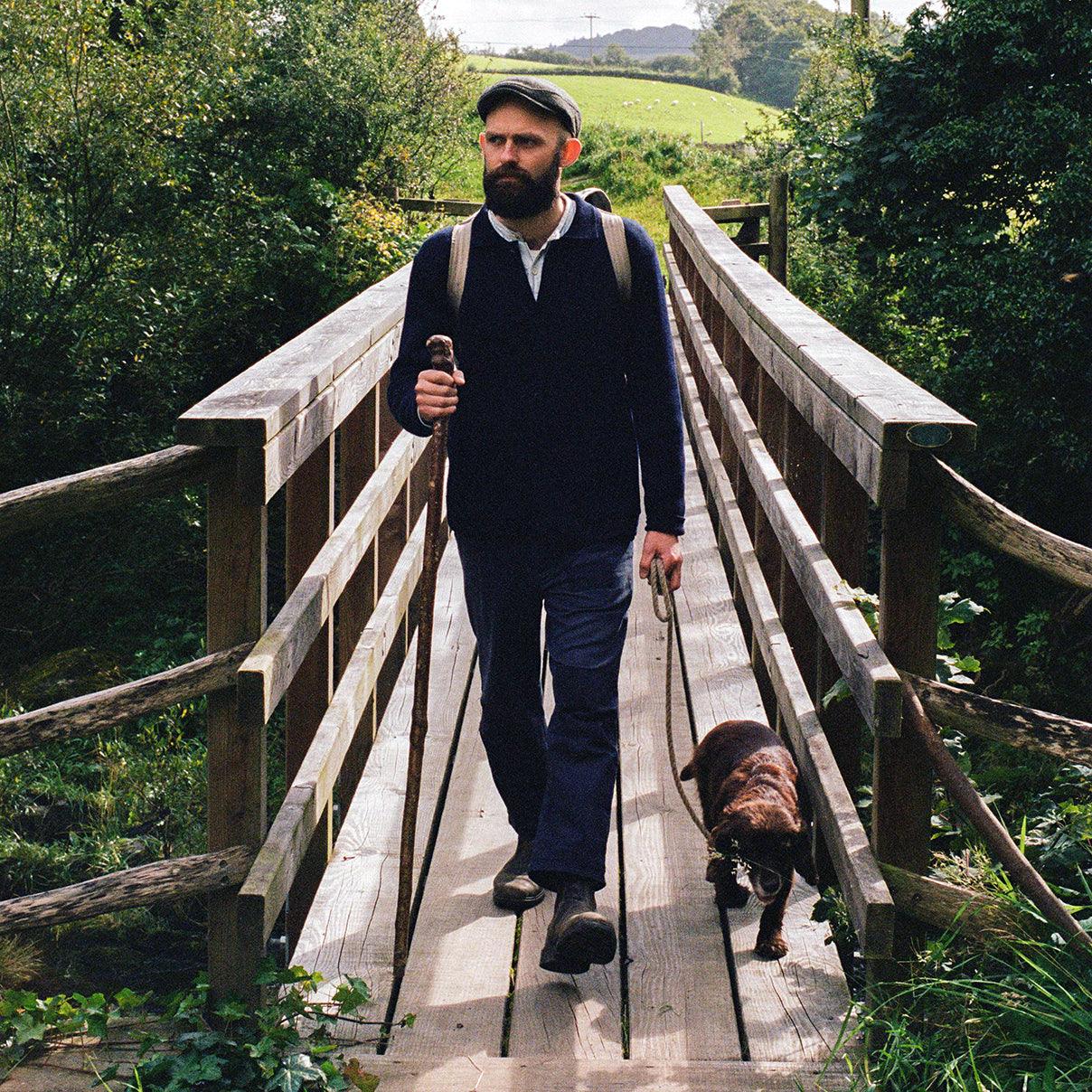 Un compagnon de labeur revêtu d'une veste de travail bleue en laine et d'un pantalon de travail assorti de la marque Champ de Manœuvres, se promenant à travers les comtés anglais avec son fidèle compagnon canin.